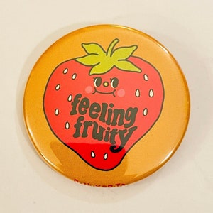 Może przedstawiać: Okrągły, pomarańczowy guzik z ilustracją czerwonej truskawki. Truskawka ma uśmiechniętą twarz i napis "feeling fruity" w kolorze czarnym.