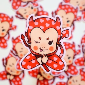 May include: A red and pink devil costume sticker with a winking baby face and a tongue sticking out. The costume has a heart pattern and horns.