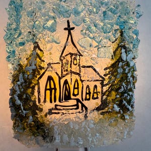 May include: A decorative night light featuring a church scene. The church is depicted with black outlines and yellow windows, set against a snowy landscape. The edges are adorned with blue and clear glass pieces, creating a textured, illuminated effect.