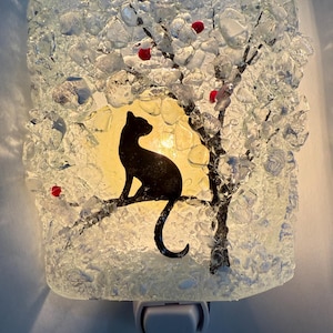 May include: A decorative night light featuring a black cat silhouette perched on a tree branch. The tree has red accents and is set against a yellow background. The light is encased in clear, textured glass.