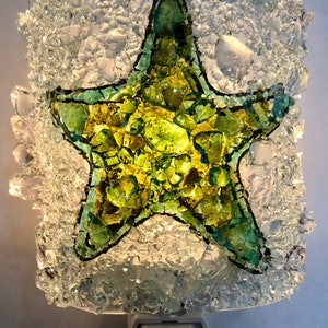 May include: A night light featuring a green and yellow glass mosaic starfish on a clear glass background. The night light has a white plug-in base with an on/off switch.
