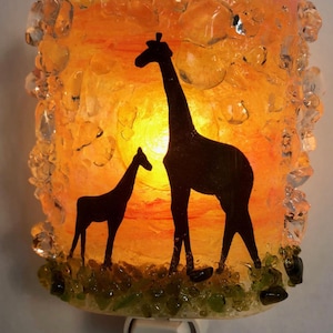 May include: A glass nightlight with a textured surface features a silhouette of a giraffe and a baby giraffe against a sunset background. The nightlight has a white plug-in base.