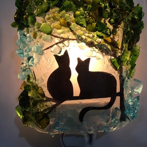May include: A nightlight featuring two black cat silhouettes under a green glass tree with blue glass accents.