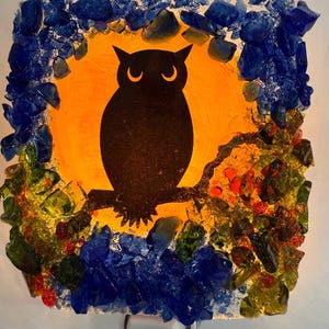 May include: A square night light featuring a black owl silhouette perched on a branch against an orange background. The edges are adorned with colorful, textured glass pieces in blue, green, yellow, and red.