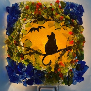May include: A stained glass nightlight with a black cat silhouette sitting on a branch in front of a yellow moon with a bat flying above. The nightlight is surrounded by blue and green glass pieces.