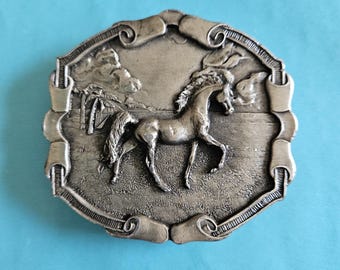 May include: Silver-toned metal belt buckle featuring a detailed relief of a horse in a landscape scene. The buckle has an ornate, scrolled border and is set against a light blue background. The horse is the central focus.