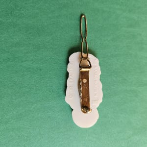 Peut inclure: Barrette &agrave; cheveux en plastique blanc avec un motif floral. Le dos de la barrette a une barre en m&eacute;tal dor&eacute; avec le texte "CROSS-OVA INC." et "MADE IN KOREA".