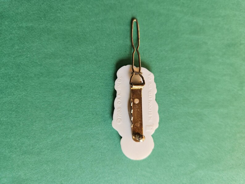 Peut inclure: Barrette &agrave; cheveux en plastique blanc avec un motif floral. Le dos de la barrette a une barre en m&eacute;tal dor&eacute; avec le texte "CROSS-OVA INC." et "MADE IN KOREA".