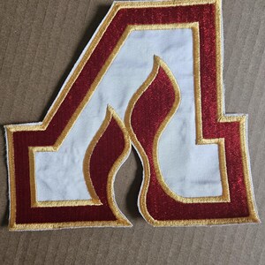 May include: A white felt patch with a red embroidered letter "A" with a flame design inside. The letter is outlined in gold thread.