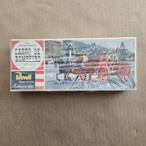 May include: A vintage model kit box for a horse-drawn fire engine. The box features a colorful illustration of the fire engine in a city setting. The text on the box reads "Carro de Bombeiro" and "Revell Authentic Kits".