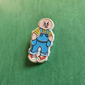 May include: A white enamel pin featuring a cartoon character wearing blue overalls, a yellow shirt, and red shoes. The character has a bald head and a friendly expression.