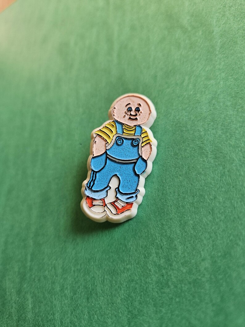 Peut inclure: Une &eacute;pingle en &eacute;mail blanc repr&eacute;sentant un personnage de dessin anim&eacute; portant une salopette bleue, une chemise jaune et des chaussures rouges. Le personnage a la t&ecirc;te chauve et une expression amicale.