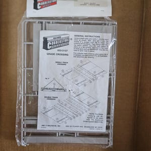 May include: A Walthers Cornerstone Series 933-3137 Grade Crossing model kit in a clear plastic bag. The kit includes gray plastic parts on a sprue and an instruction sheet with diagrams for single and double track crossing assembly configurations.