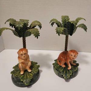 May include: Two ceramic monkey figurines, each perched beneath a miniature palm tree. The monkeys are brown with detailed facial features, sitting on green leaf-shaped bases. The palm trees have green fronds and brown trunks.