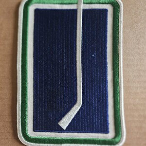 May include: Embroidered patch featuring a hockey stick and a rectangular design. The patch has a navy blue background with a white hockey stick. The border is green, white, and green.