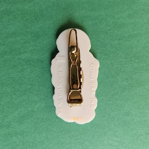 Peut inclure: Barrette &agrave; cheveux en plastique blanc avec un fermoir en m&eacute;tal dor&eacute;. La barrette pr&eacute;sente un motif floral et le texte "GREET O'MAL INC. HELDON LTD. W.Y. CA."