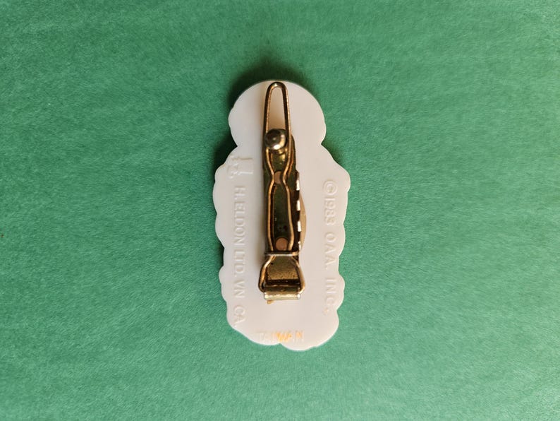 Peut inclure: Barrette &agrave; cheveux en plastique blanc avec un fermoir en m&eacute;tal dor&eacute;. La barrette pr&eacute;sente un motif floral et le texte "GREET O'MAL INC. HELDON LTD. W.Y. CA."