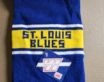 May include: Blue and yellow knitted scarf with the text "ST. LOUIS BLUES" in white. The scarf has a white tag with the text "Wear-A-Knit" and a logo.