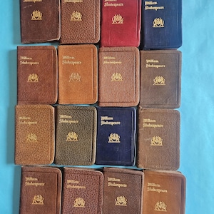 May include: A collection of miniature books with embossed gold lettering and a crest. The books are in various colors, including brown, red, and blue. The title "William Shakespeare" is visible on the covers.