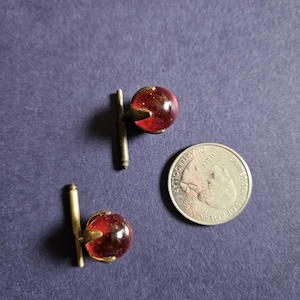 May include: A pair of cufflinks featuring round, translucent red glass spheres held by brass-toned metal prongs and T-bar fasteners. A United States quarter coin is placed next to the cufflinks to provide a clear sense of scale for the items.