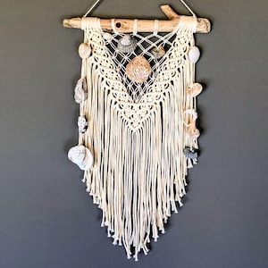 Könnte beinhalten: Ein Wandbehang aus cremefarbenem Makramee-Garn, das in einem V-förmigen Muster geknotet und an einem Stück natürlichem Treibholz befestigt ist. Das Design ist mit verschiedenen Muscheln unterschiedlicher Form, Größe und Textur verziert.