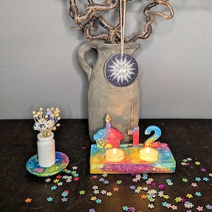 Könnte beinhalten: Ein mehrfarbiger Kerzenhalter aus Kunstharz mit der Zahl 12 und einer Cupcake-Form, der zwei brennende Teelichter enthält. Daneben steht eine kleine weiße Vase mit Trockenblumen auf einer passenden Untertasse, umgeben von buntem Blumenkonfetti.