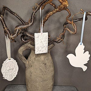 Könnte beinhalten: Drei weiße Keramik-Anhänger hängen an einem Ast. Die Anhänger sind in Form eines Vogels, eines Herzens mit einem Vogel darin und eines Herzens mit einem Blumenmuster. Der Ast ist braun und hat eine raue Textur.