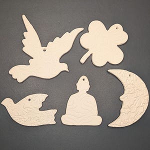 Könnte beinhalten: Fünf Holzornamente in verschiedenen Formen, darunter eine Taube, ein Kleeblatt, ein Halbmond, eine meditierende Figur und ein Vogel. Die Ornamente sind alle weiß und haben eine strukturierte Oberfläche.