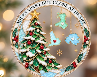 Puede incluir: Un adorno navideño circular con un árbol de Navidad verde, adornos rojos y blancos y una estrella dorada en la punta. El texto MILES APART BUT CLOSE AT HEART aparece en la parte superior. Tres formas de mapas cuelgan del marco con corazones.