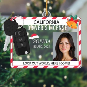 May include: A red and white striped Christmas ornament shaped like a California driver's license. The ornament features a photo of a woman with a Santa hat, the name "Sophia", and the text "Issued: 2024". The ornament also includes a key and a key fob.