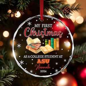 May include: A clear, faceted Christmas ornament with a red ribbon. The ornament features the text "MY FIRST Christmas" in red script, along with illustrations of books and snowflakes. Below the text is "AS A COLLEGE STUDENT AT ASU Hannah 2024".