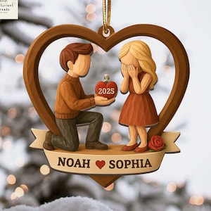 May include: A wooden ornament depicting a marriage proposal scene. A man kneels, offering a heart-shaped object with "2025" on it to a woman. The ornament is heart-shaped with the names "NOAH" and "SOPHIA" on a banner.