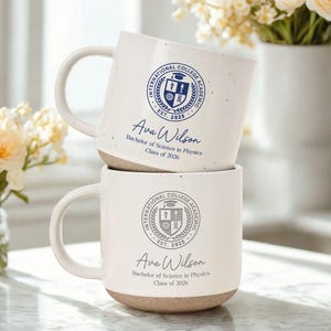 May include: Two stacked white ceramic mugs with a speckled finish. Each mug features a blue college emblem, the name "Ava Wilson", and "Bachelor of Science in Physics, Class of 2026". The mugs have a beige base and a curved handle.