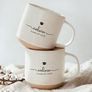 May include: Two stacked off-white ceramic mugs with a light brown base. Each mug has a curved handle and features the name "calvin" in a script font, a graduation cap icon, and the text "CLASS OF 2026" in black.