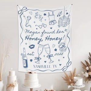 May include: White tapestry with blue line art illustrations of champagne glasses, a disco ball, and the text "Megan found her Honey, Honey!" with "Nashville, TN" and the year 2025. The tapestry is hanging above a table with cakes.
