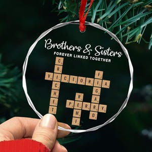 May include: A clear, round Christmas ornament with a red ribbon. The ornament features the text "Brothers & Sisters FOREVER LINKED TOGETHER." Scrabble-style tiles spell out names. The ornament is held by a person.