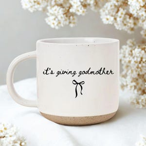 May include: A white ceramic mug with a brown base and a curved handle. The mug features the phrase "it's giving godmother" in black script, along with a black bow graphic. The background includes white flowers.