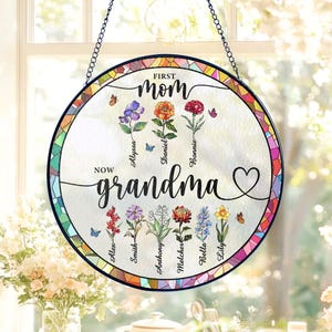 May include: Round stained glass window art with the words "First Mom Now Grandma" and names of family members. The design features colorful flowers, butterflies, and a heart. A decorative piece for a home.