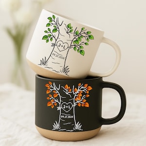 May include: Two ceramic mugs stacked vertically. The top mug is off-white with a green tree illustration, a heart containing J+L, and the date 09.27.2026. The bottom mug is black with an orange tree illustration, a heart with J+L, and 09.27.2026.