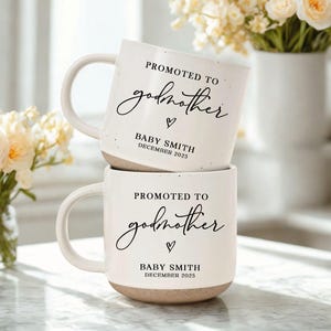 May include: Two stacked white ceramic mugs with a speckled finish and a beige base. Each mug has the text "PROMOTED TO godmother" in a script font, a heart symbol, and "BABY SMITH DECEMBER 2025". The mugs are near a vase of flowers.