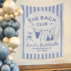 May include: A white and blue striped tapestry with the text "THE BACH CLUB EST 2026" in blue. The tapestry features a drawing of a cocktail and lingerie hanging on a clothesline. The text "Sarah's Bachelorette Miami Beach, FL EST. 2026" is also included.