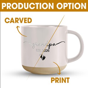 May include: White ceramic mug with a beige base, featuring the word "grandpa" in a script font, the year "EST. 2026", and baby footprints. The mug has a curved handle and is labeled with "CARVED" and "PRINT".