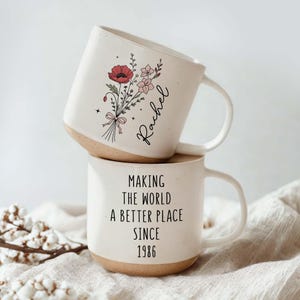 May include: Two stacked white ceramic mugs. The top mug features a floral design with a red poppy and the name "Rachel". The bottom mug has the text "MAKING THE WORLD A BETTER PLACE SINCE 1986". Both mugs have a tan base.