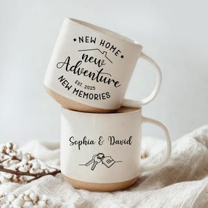 May include: Two stacked ceramic mugs with a cream-colored exterior and a tan base. The top mug has the text "NEW HOME, new Adventure, NEW MEMORIES, Est. 2025". The bottom mug says "Sophia & David" with a key and house graphic.