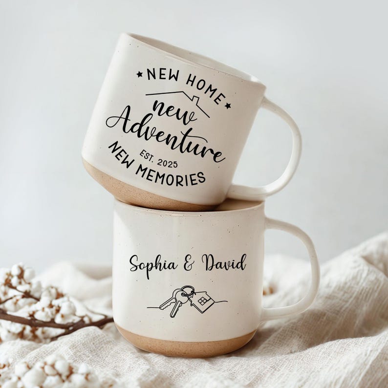 May include: Two stacked ceramic mugs with a cream-colored exterior and a tan base. The top mug has the text "NEW HOME, new Adventure, NEW MEMORIES, Est. 2025". The bottom mug says "Sophia & David" with a key and house graphic.
