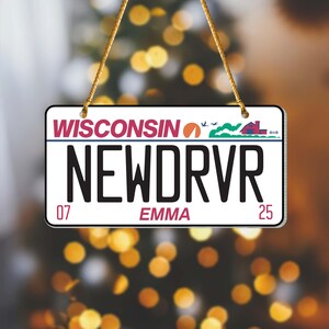 Puede incluir: Un letrero rectangular que se asemeja a una placa de matr&iacute;cula de Wisconsin, con el texto "WISCONSIN" en la parte superior. La placa dice "NEWDRVR" en letras negras grandes, con "EMMA" y los n&uacute;meros "07" y "25" debajo. El letrero est&aacute; suspendido por una cuerda dorada.