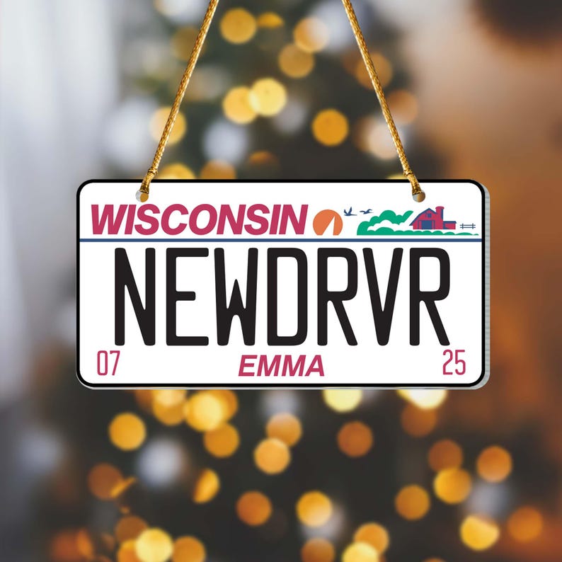 Puede incluir: Un letrero rectangular que se asemeja a una placa de matr&iacute;cula de Wisconsin, con el texto "WISCONSIN" en la parte superior. La placa dice "NEWDRVR" en letras negras grandes, con "EMMA" y los n&uacute;meros "07" y "25" debajo. El letrero est&aacute; suspendido por una cuerda dorada.