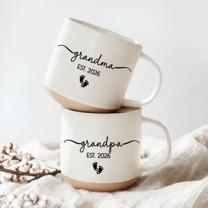 May include: Two white ceramic mugs stacked on top of each other. The mugs have a speckled finish with a tan base. The top mug says "grandma EST. 2026" with a footprint graphic. The bottom mug says "grandpa EST. 2026" with a footprint graphic.