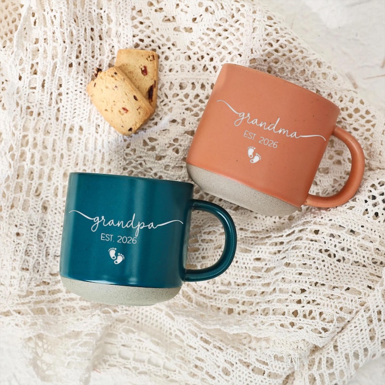 May include: Two ceramic mugs, one teal and one peach, with gray bottoms and curved handles. The teal mug says "grandpa EST. 2026" and the peach mug says "grandma EST. 2026" in white script. Both mugs have a footprint graphic. A few cookies are in the background.