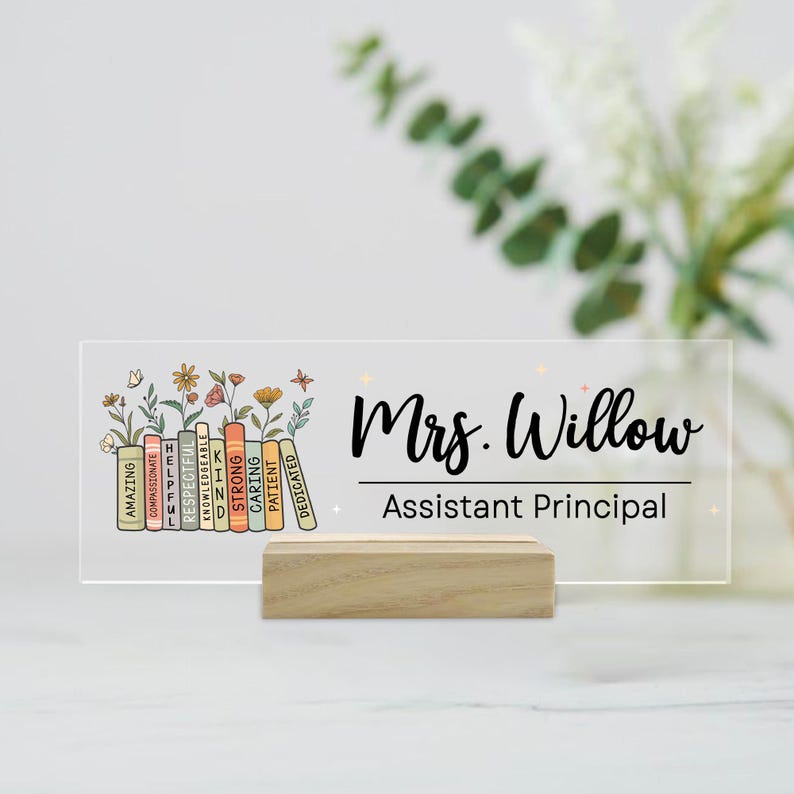 May include: A clear acrylic desk sign with the name "Mrs. Willow" and the title "Assistant Principal" in black script. The sign features a floral design and a stack of colorful books with positive adjectives. It is displayed on a wooden base.